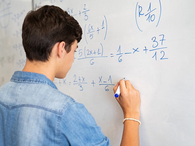 ack view of high school student solving math problem on whiteboard in classroom.