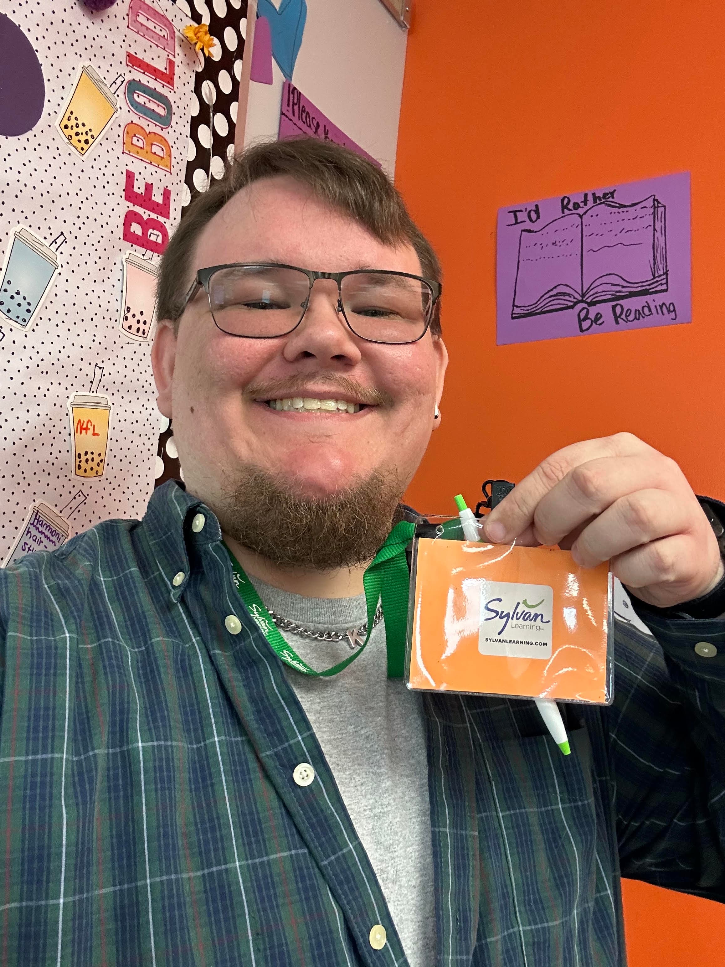 Smiling man in glasses in front of a classroom wall holding a Sylvan lanyard