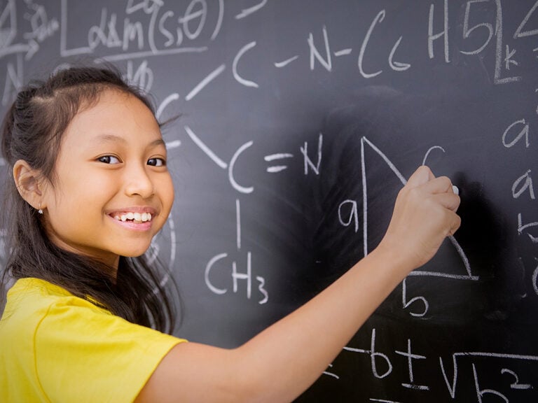 Female elementary student smiling at the camera while writing mathematical formula on the blackboard