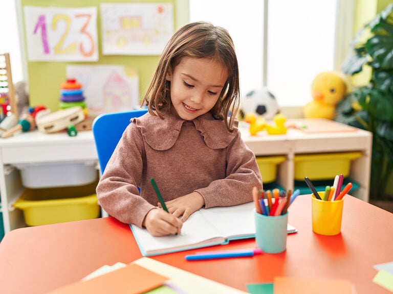 Little girl preschool student sitting on table writing on notebook at kindergarten
