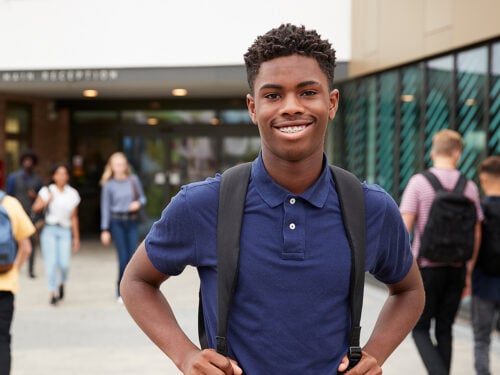 Portrait Of Smiling Male High School Student Outside College Building With Other Teenage Students In Background